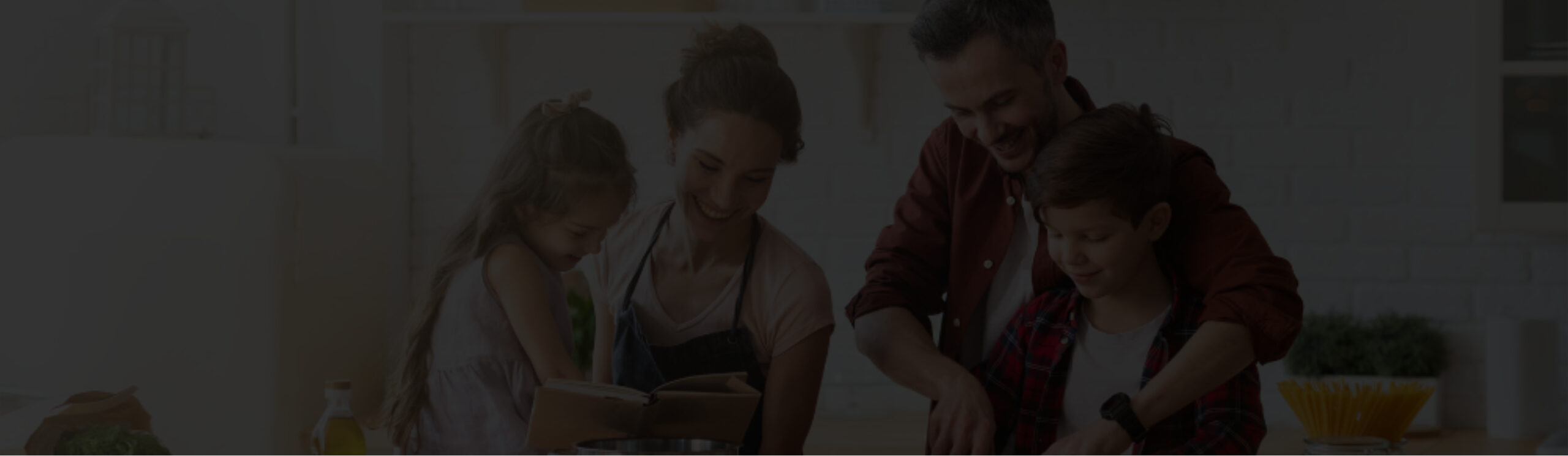 A family of four stands together in a kitchen, with the adults assisting the children in cooking or preparing food.