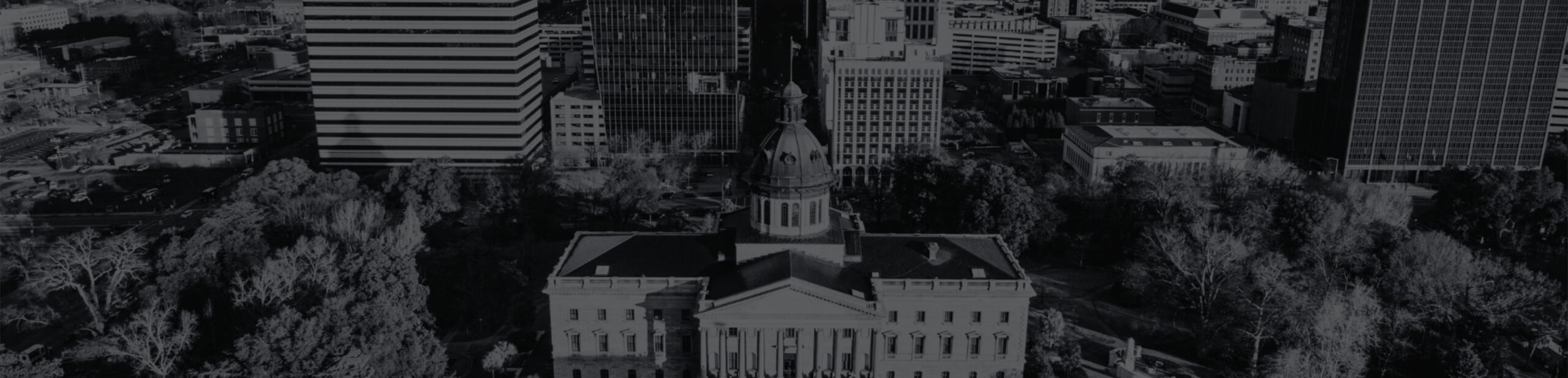 Aerial view of a government building with a dome, surrounded by trees and modern office buildings in a cityscape.