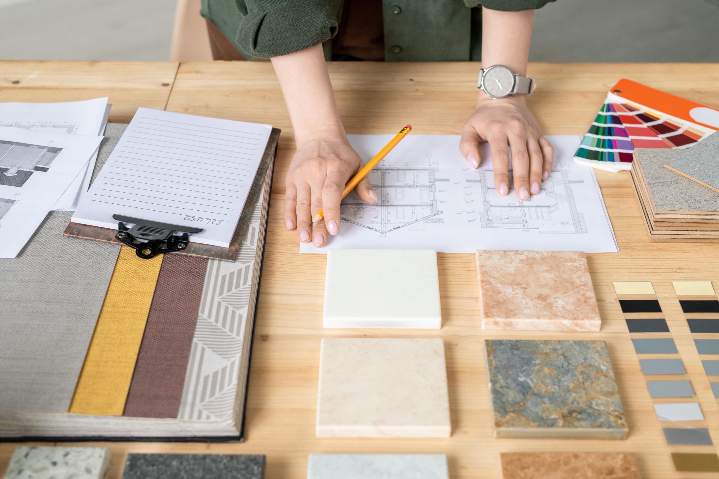 Person reviewing architectural plans at a table with tiles, fabric swatches, color samples, and design materials laid out.
