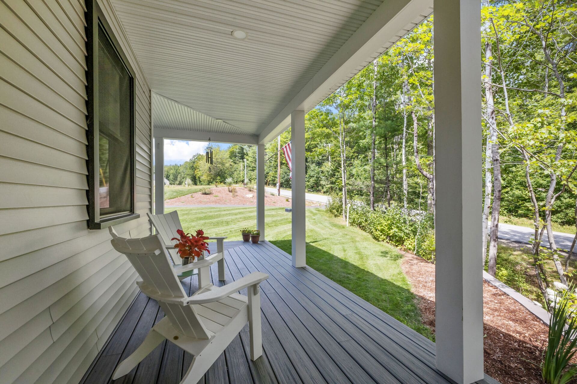 A white wooden chair with a small table and potted plants sits on a covered porch overlooking a green yard lined with trees and an American flag in the background.