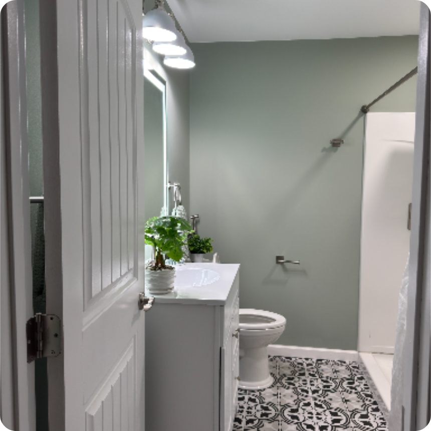 A small bathroom with a light green wall, patterned floor tiles, a white vanity with a potted plant, a toilet, and a shower with a white curtain.