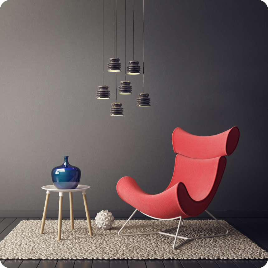 A modern interior with a red accent chair, a small round table holding a blue vase, a decorative object on a textured rug, and pendant lights against a dark wall.