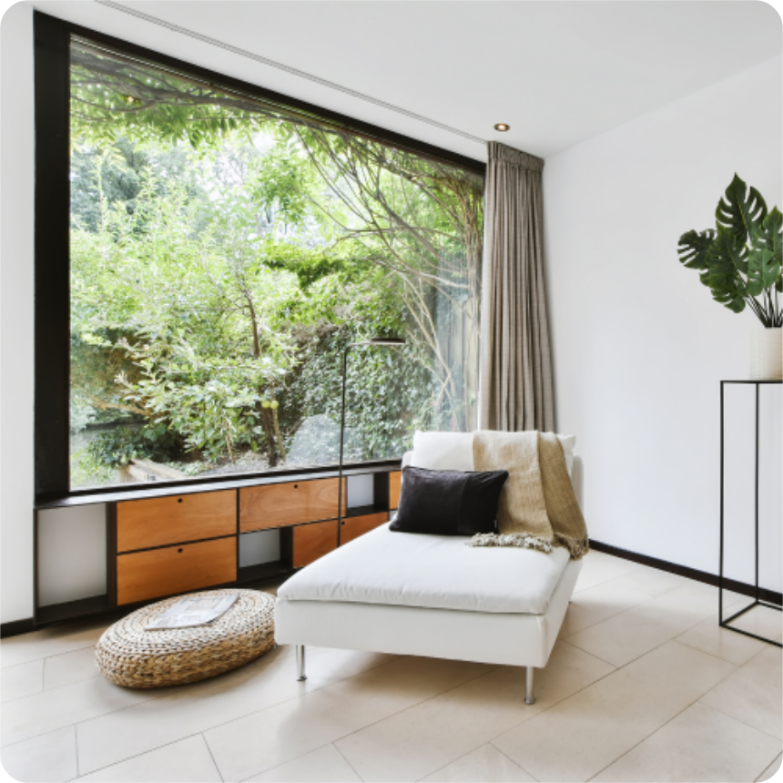 Modern minimalist living space with a white chaise lounge, large window overlooking greenery, a woven pouf, and light wooden storage cabinet below the window.