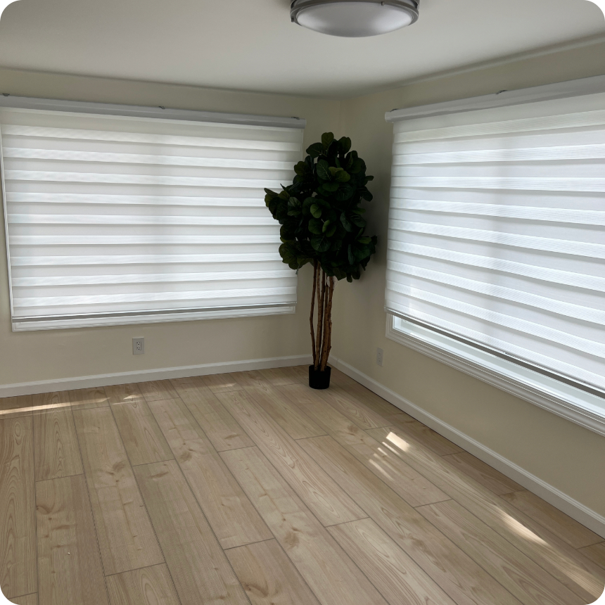 A bright, empty room with light wood flooring, two large windows with white blinds, and a potted artificial plant in the corner.