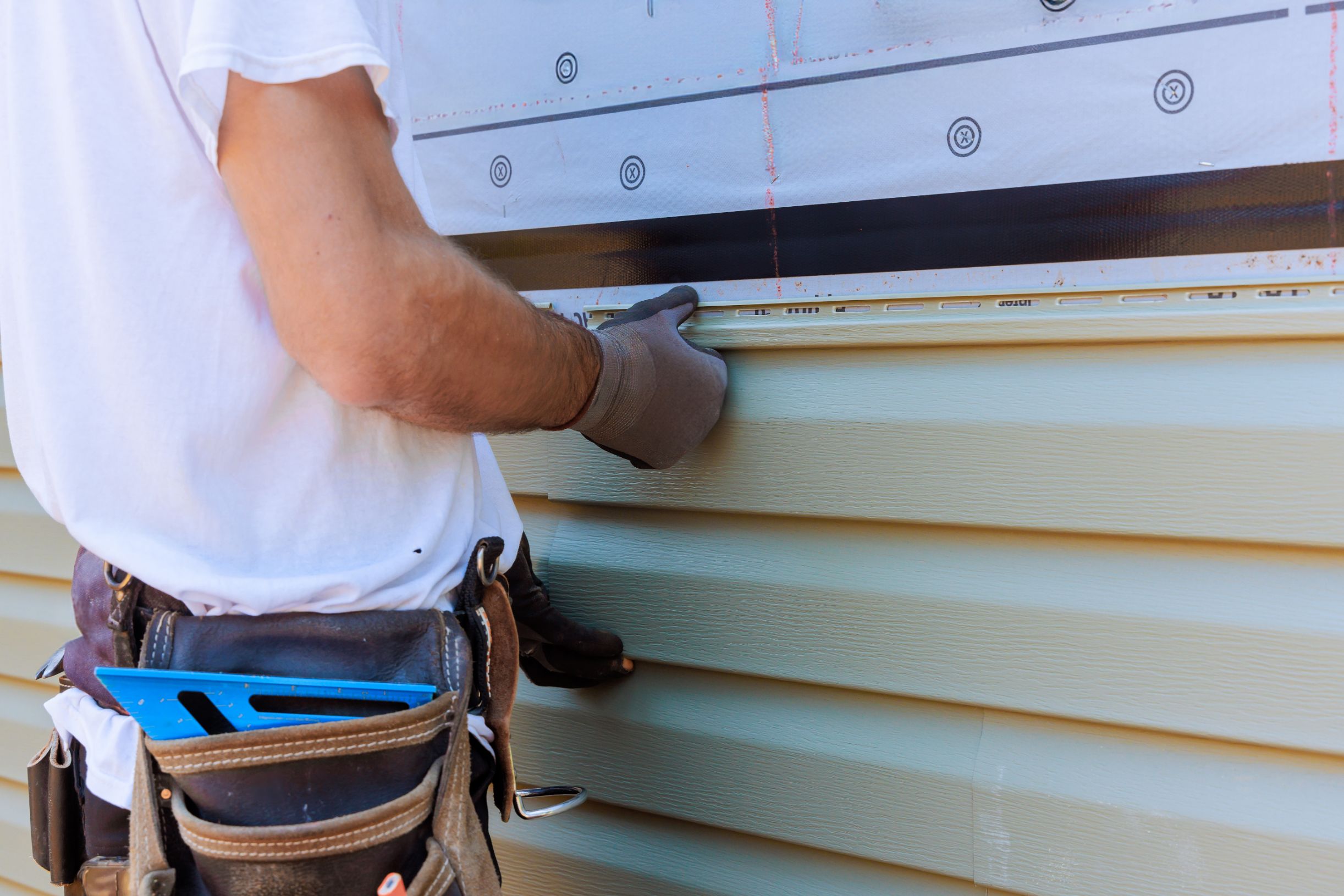 A person wearing gloves installs beige vinyl siding on a house exterior, using tools stored in a brown leather tool belt.