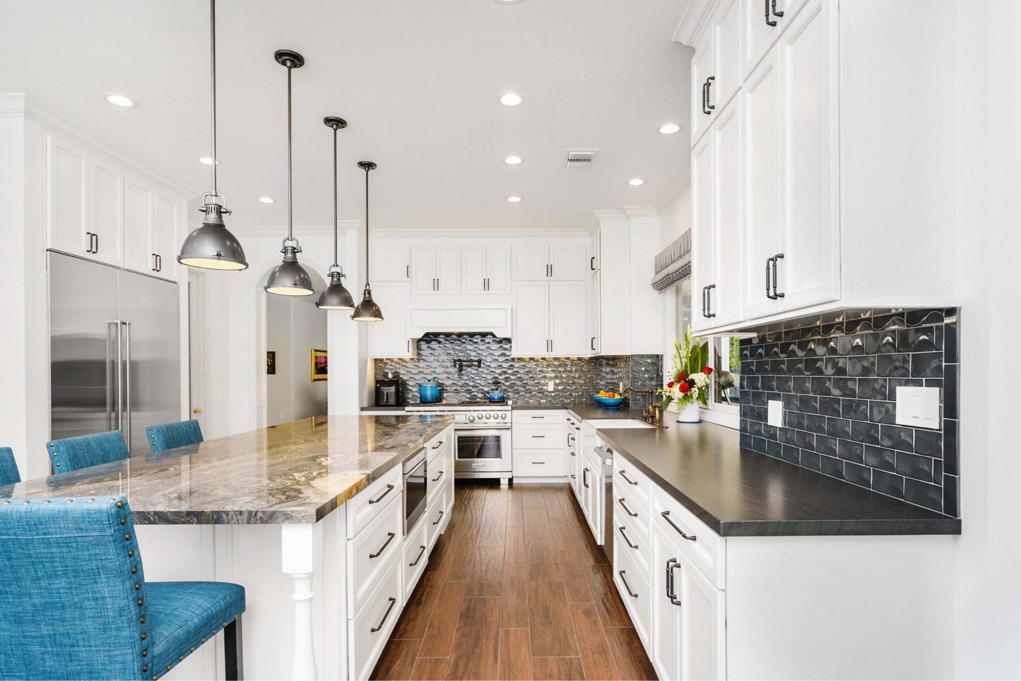 Modern kitchen with white cabinets, black tile backsplash, stainless steel appliances, marble island, pendant lights, and blue upholstered bar stools on wood flooring.