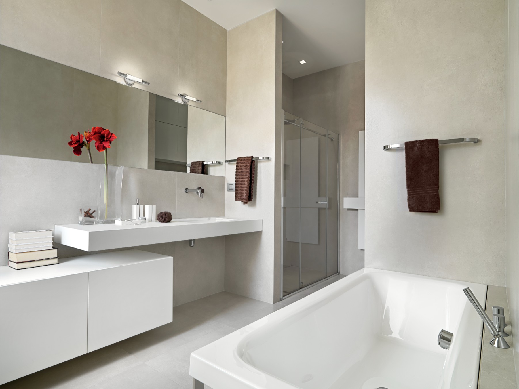 Modern bathroom with a white bathtub, floating vanity with double sink, large mirrors, glass shower, and red flowers on the counter. Brown towels and neutral tones throughout.