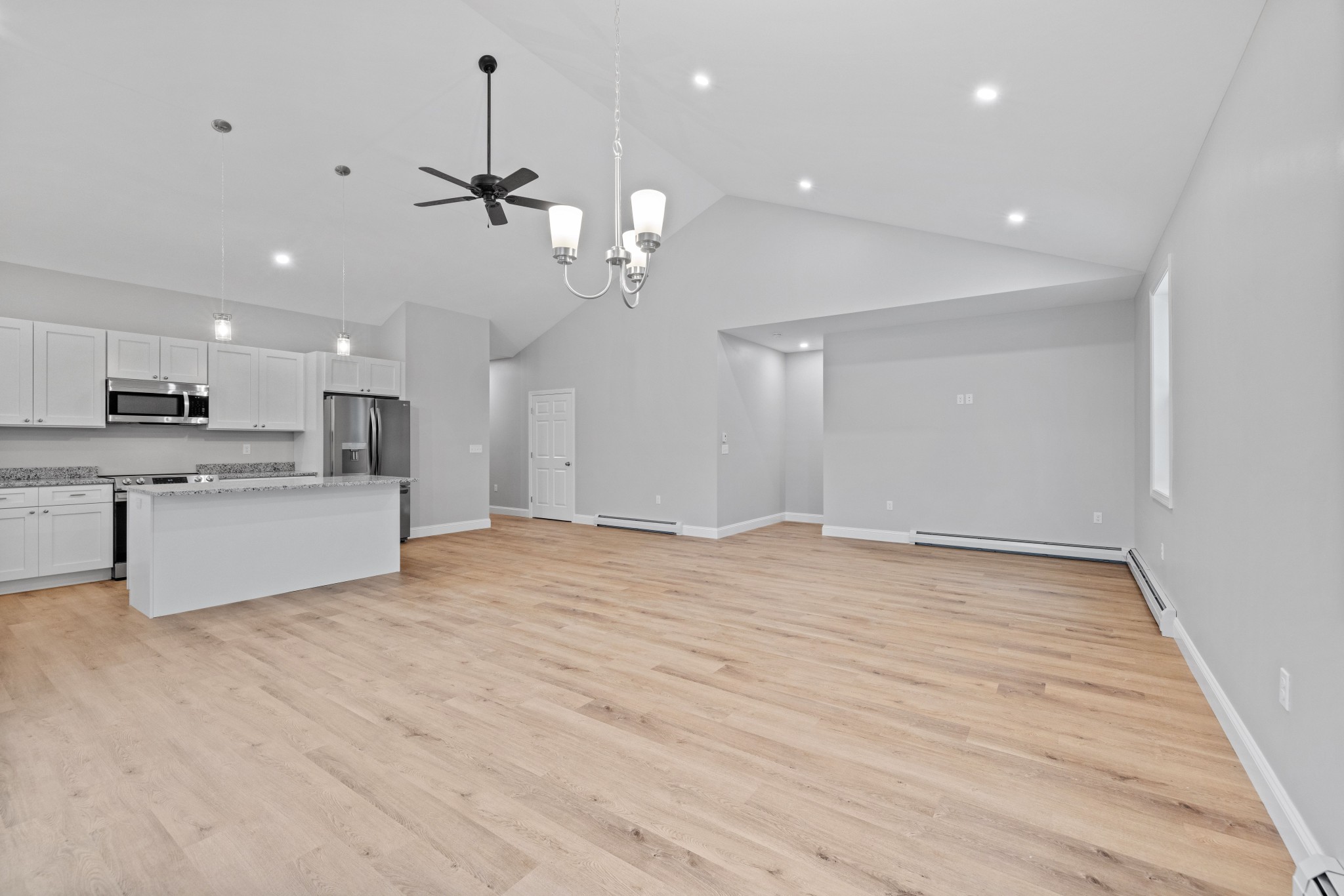 Spacious open-plan living area with light wood flooring, white walls, a modern kitchen with stainless steel appliances, island, and ceiling lights, featuring large windows and vaulted ceiling.
