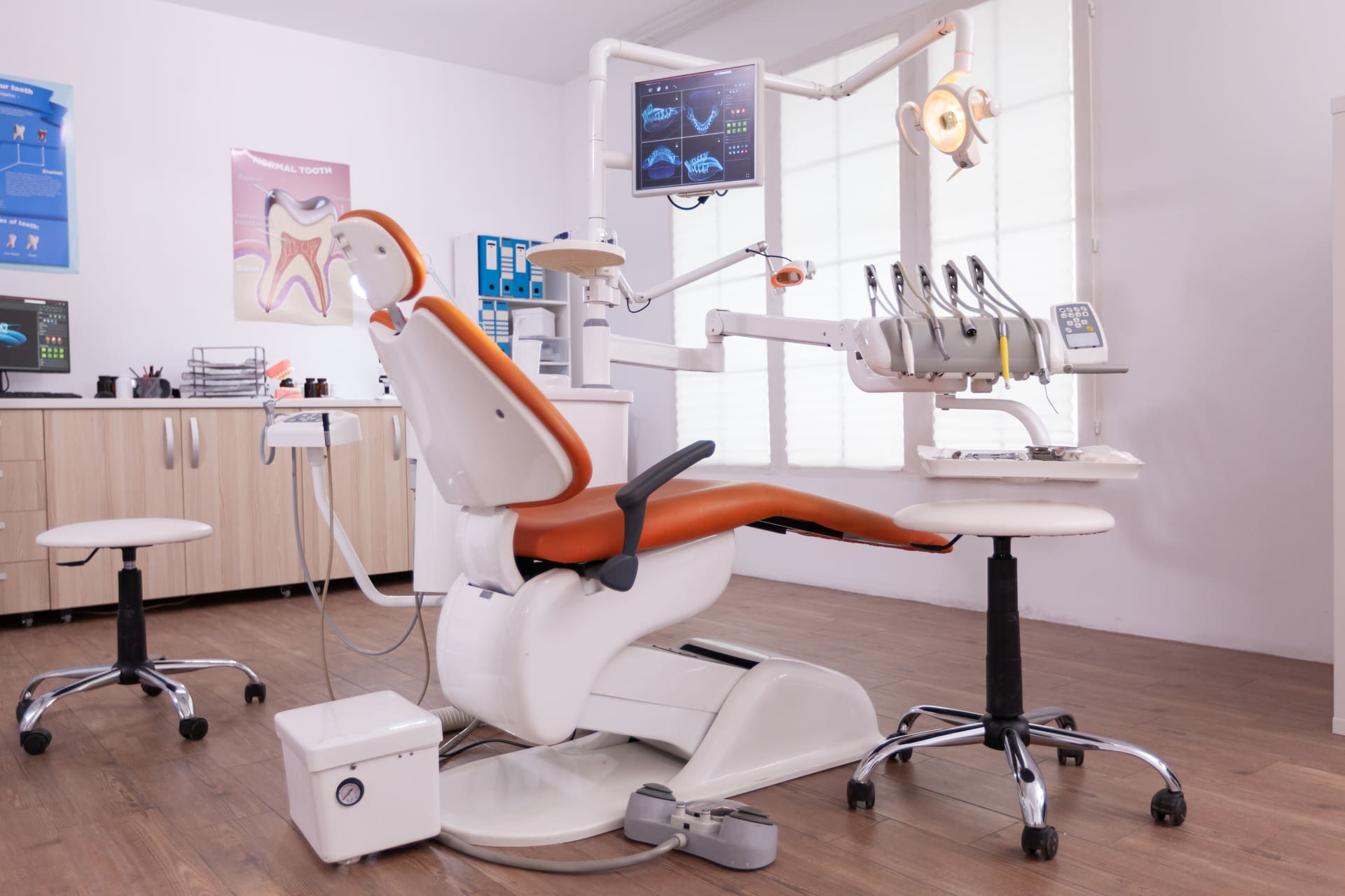Modern dental clinic room with an orange dental chair, dental instruments, monitor, and bright lighting on a wooden floor.