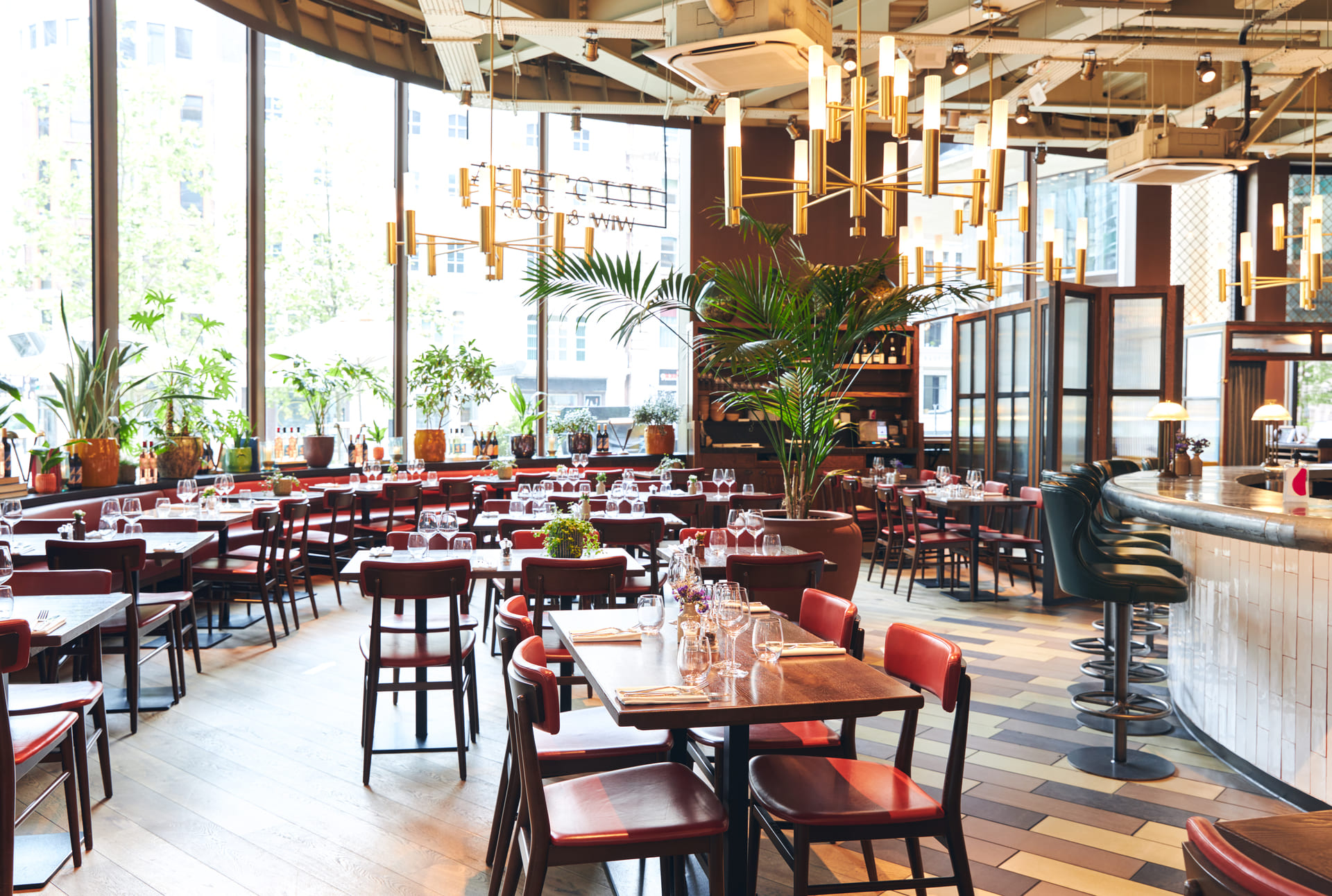 Modern restaurant interior with red chairs, wooden tables set with glasses and cutlery, large windows, plants, and a curved bar with barstools.