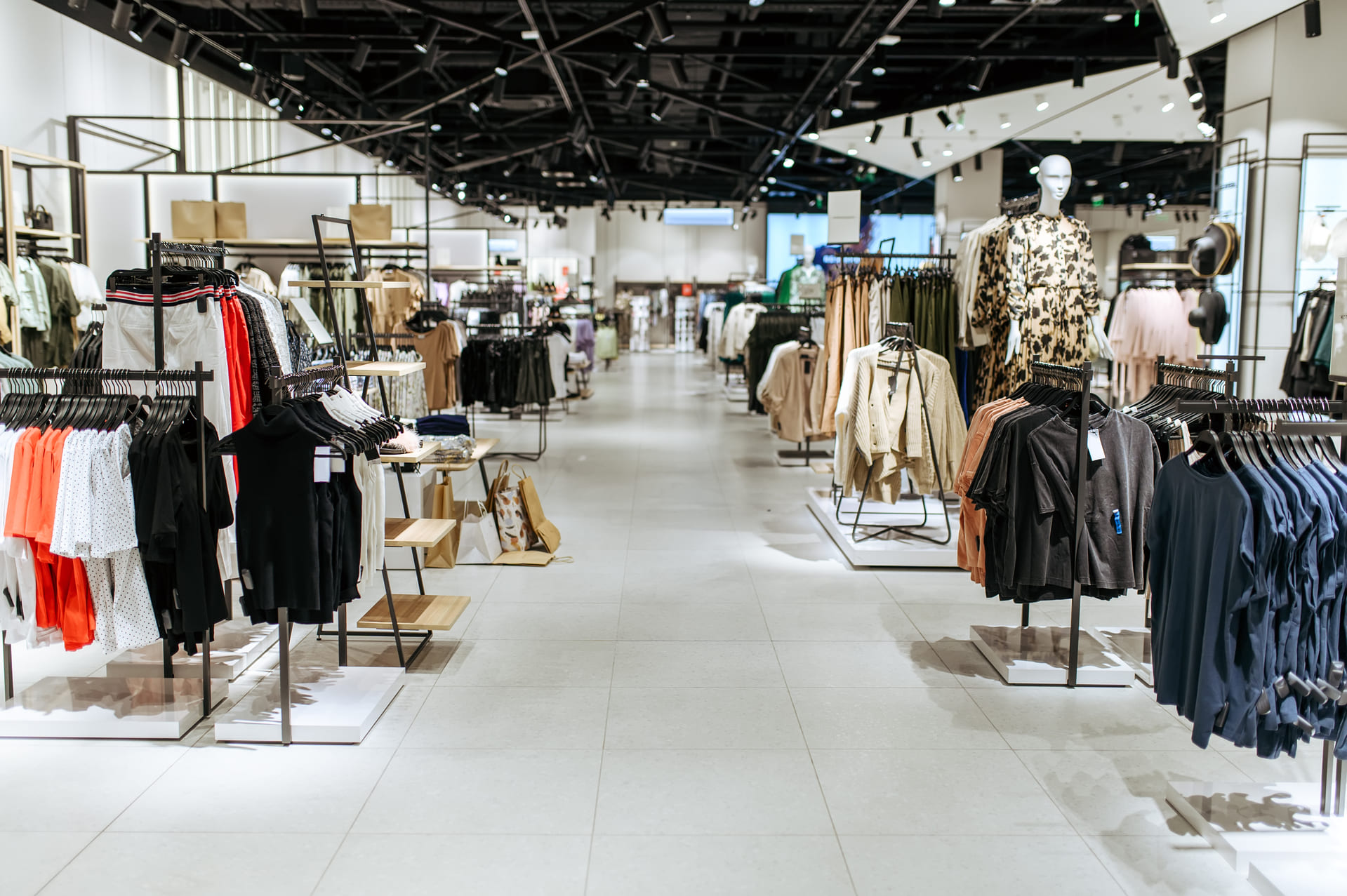 Clothing racks and mannequins are displayed in a spacious, well-lit retail clothing store with various garments organized by type.
